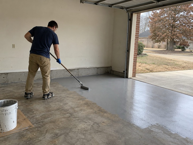 Installer in spiked shoes rolling a gray base coat across a garage floor